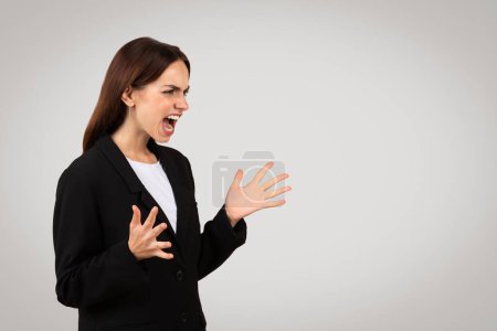 Photo for Angry millennial european businesswoman shouting with a fierce expression, her hands open in a defensive or confrontational manner, wearing a classic black business suit, studio - Royalty Free Image