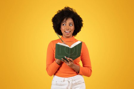 Foto de Mujer joven curiosa con el pelo rizado, cuello alto naranja, con un libro verde y un lápiz en la cara, con una expresión curiosa, sobre un fondo amarillo - Imagen libre de derechos