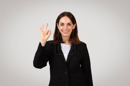 Foto de Alegre millennial empresaria europea en un traje negro inteligente haciendo un signo positivo de mano bien, que transmite la aprobación o la satisfacción, en un fondo neutral, estudio - Imagen libre de derechos