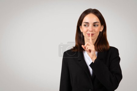 Photo for Playful millennial european businesswoman with a mischievous grin placing her finger over lips in a shush gesture, signaling a need for secrecy or quiet, in a professional black suit - Royalty Free Image