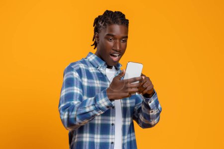 Foto de Gran aplicación. Hombre negro joven asombrado mirando la pantalla del teléfono inteligente con emoción, reaccionando emocionalmente a la nueva aplicación, de pie con la boca abierta aislada en el fondo amarillo, espacio de copia - Imagen libre de derechos