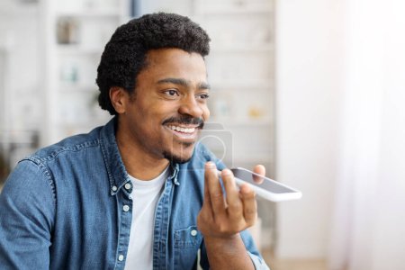 Téléchargez les photos : Un jeune homme noir souriant enregistre un message vocal sur son smartphone à la maison, un homme afro-américain millénaire utilisant un téléphone portable moderne, naviguant dans un assistant virtuel ou parlant avec un haut-parleur - en image libre de droit