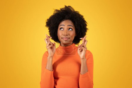Téléchargez les photos : Espérant jeune femme afro-américaine croise les doigts pour la bonne chance, les yeux regardant vers le haut avec un sourire plein de vœux, portant un pull orange sur un fond jaune vif - en image libre de droit