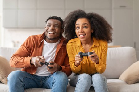 Téléchargez les photos : Joyeux couple millénariste noir jouant à des jeux vidéo sur console de manette à la maison, profitant de loisirs domestiques et de divertissement numérique, mettant en valeur le plaisir de leur activité de passe-temps partagée - en image libre de droit