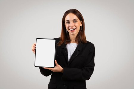 Foto de Una alegre mujer europea milenaria en traje de negocios muestra una tableta con una pantalla en blanco, perfecta para maquetas o mostrar aplicaciones y sitios web, aislados en un fondo gris, estudio - Imagen libre de derechos