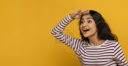 Photo for Curious pretty young hindu woman in casual holding hand next to forehead and looking at blank copy space for text or advertisement, posing isolated on yellow studio background, web-banner - Royalty Free Image