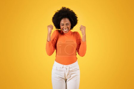 Photo for Exuberant young woman celebrates with a triumphant expression, clenching her fists in victory, donned in a vibrant orange turtleneck and white pants, against a yellow background - Royalty Free Image