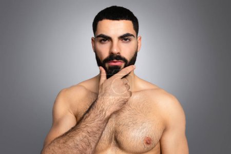 Tips for healthy beard concept. Handsome athletic naked brunette middle eastern young man touching his thick beard and looking at camera, isolated on grey studio background, closeup