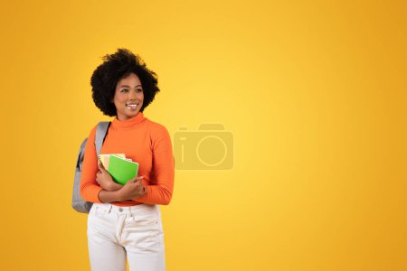 Téléchargez les photos : Joyeux jeune étudiant avec une coiffure naturelle se dresse sur un fond jaune vif, tenant des carnets colorés et portant un col roulé orange vif, un pantalon blanc et un sac à dos gris - en image libre de droit