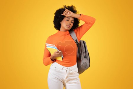 Téléchargez les photos : Jeune étudiante bouleversée se sentant stressée, tenant son front avec des cahiers à la main, vêtue d'un haut orange et d'un jean blanc avec un sac à dos gris, sur un fond jaune - en image libre de droit