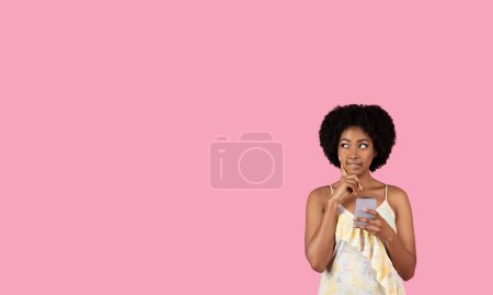 Téléchargez les photos : Une jeune femme africaine américaine réfléchie aux cheveux bouclés tient un smartphone et regarde pensivement sur un fond rose uni, incarnant la contemplation et la communication moderne - en image libre de droit