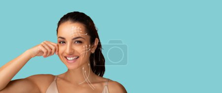 Photo for Beautiful young woman with clean fresh skin touching her chin, enjoying antiaging massage. Cheerful pretty lady with lifting arrows on her face, blue studio background, panorama with copy space - Royalty Free Image