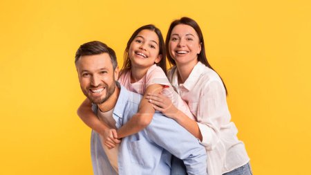 Téléchargez les photos : Portrait de famille de parents joyeux et de leur petite fille embrassant, debout en studio sur fond jaune, câlinant et s'amusant en posant ensemble, panorama - en image libre de droit