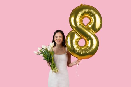 Photo for Smiling woman celebrating Womens Day with white tulips and golden balloon shaped as number 8, symbolizing March 8th, against cheerful pink background - Royalty Free Image
