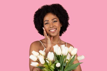 Photo for A joyful millennial African American woman with a radiant smile and curly hair, gently touching her cheek, holds a bouquet of fresh white tulips against a pink background - Royalty Free Image