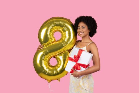 Photo for Smiling millennial african american woman with a natural afro hairstyle holding a golden number 8 balloon and a white gift box with a red ribbon against a pink backdrop, studio - Royalty Free Image