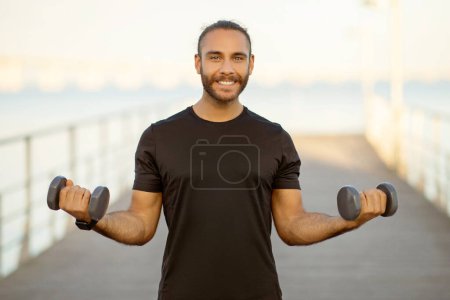 Photo for Motivated man strengthens his arms muscles lifting weights on sea pier outdoors. Guy exercising with dumbbells smiling to camera. Concept of fitness workout and healthy lifestyle - Royalty Free Image