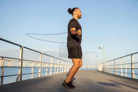 Sporty man by the sea engages in morning aerobics jumping with skipping rope in activewear outdoor, side view, full length shot. Fitness motivation, showcasing healthy and active lifestyle