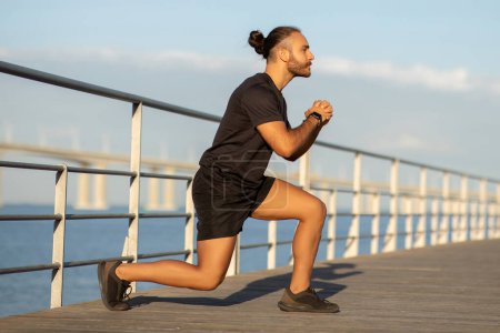 Photo for Outdoor fitness. Side view of motivated young man in sportswear demonstrating a lunge deep exercise, promoting healthy and active lifestyle by the sea pier. Workouts and wellbeing - Royalty Free Image