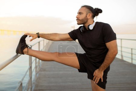 Photo for Fit European guy in sportswear practices a standing leg stretch outside, promoting healthy lifestyle, workout motivation for muscles flexibility. Sport and wellness by the sea - Royalty Free Image