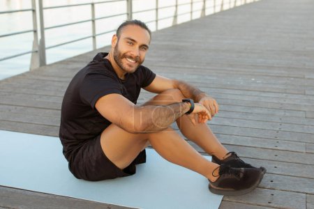 Photo for Fitness joy. Happy sporty guy in activewear takes a break on a pier, sitting on gym mat outside, radiating wellbeing and happiness as he smiles at camera. Concept of healthy lifestyle and workout - Royalty Free Image