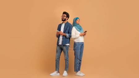 Photo for Portrait Of Young Muslim Couple With Smartphones Looking At Copy Space With Interest, Smiling Arabic Man And Woman In Hijab Holding Mobile Phones, Standing Back To Back On Beige Studio Background - Royalty Free Image