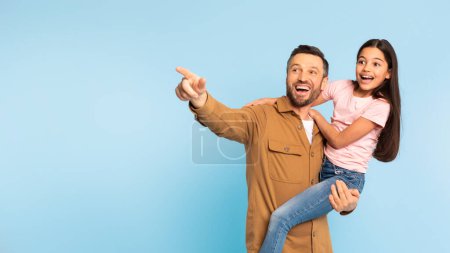 Téléchargez les photos : Offre familiale. Happy Man tenant sa fille enfant dans les bras et pointant du doigt à part, montrant l'espace de copie à l'enfant excité sur fond bleu studio, Panorama. Look There Concept - en image libre de droit