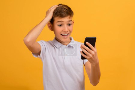 Téléchargez les photos : Incroyable adolescent garçon regardant l'écran du smartphone et touchant la tête, adolescent garçon tenant téléphone mobile avec expression de surprise et d'excitation, debout isolé sur fond de studio jaune - en image libre de droit