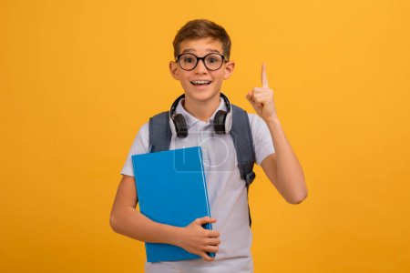 Photo for Excited Teen Boy Wearing Eyeglasses Having Idea, Raising Finger Up, Clever Teenage Male Child Holding Books, Ready To Answer A Question, Standing On Yellow Studio Background, Copy Space - Royalty Free Image