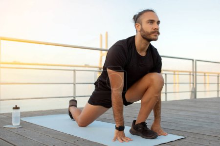 Photo for Sportsman practicing aerobics by the sea doing legs stretch outdoor, emphasizing a healthy and flexible lifestyle in summer. Guy in fitness outfit having workout on mat outside - Royalty Free Image