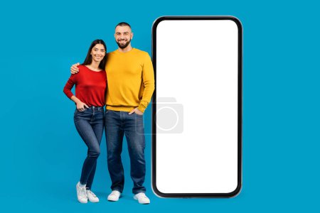 Photo for Happy young couple standing next to giant smartphone with blank white screen, smiling man and woman promoting mobile application or website, advertising your product, blue studio background, mockup - Royalty Free Image