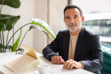 Photo for Mature businessman with a beard and mustache smiles gently while sitting in a modern office with a cup of coffee and an open notebook on a white table, with large green plants in the background - Royalty Free Image