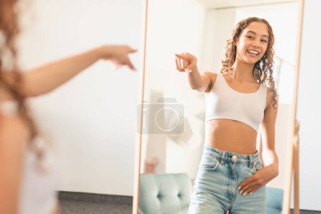 Téléchargez les photos : Adolescente caucasienne confiante pointant vers son reflet dans le miroir intérieur, favorisant l'amour-propre et l'image corporelle positive, souriant à elle-même tout en posant à la maison. Beauté et bien-être - en image libre de droit