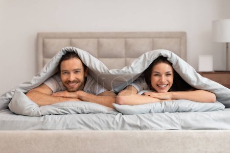 Joyful couple lies in bed under soft duvet, their faces framed by the sheets as they share playful and intimate moment, exuding happiness and comfort