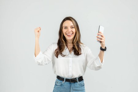 Foto de Mujer joven exuberante con un reloj inteligente, levantando triunfalmente un puño mientras sostiene un teléfono inteligente en la otra mano, con una blusa blanca y jeans, exudando positividad sobre un fondo gris - Imagen libre de derechos