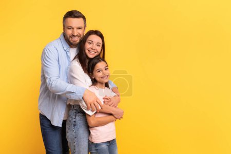 Photo for Family Offer. Happy Caucasian Man Hugging His Wife And Kid Daughter Standing Together On Yellow Backdrop, Posing Near Free Space For Text, Advertising Nice Deal Or Shopping Sale - Royalty Free Image