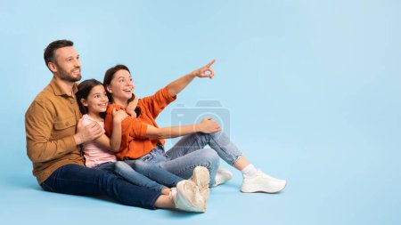 Photo for Family sitting close on floor joyfully pointing to blank space ideal for content advertisement, directing attention to free place for ads and offers, posing on blue studio background, panorama - Royalty Free Image
