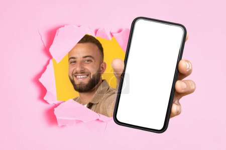 Foto de Un hombre barbudo sonriente con una camisa de pana asoma a través de un papel rosa roto, alegremente presentando un teléfono inteligente con una pantalla en blanco en la mano. Recomendación sitio web, aplicación, buenas noticias, ganar y la tecnología - Imagen libre de derechos