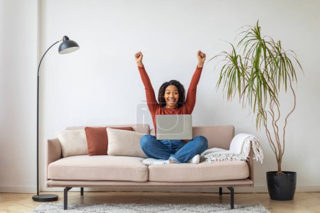Foto de Buenas noticias. Mujer negra joven y feliz celebrando el éxito con el ordenador portátil mientras se sienta en el sofá en casa, la mujer afroamericana llena de alegría levantando puños y exclamando con emoción, Copiar el espacio - Imagen libre de derechos