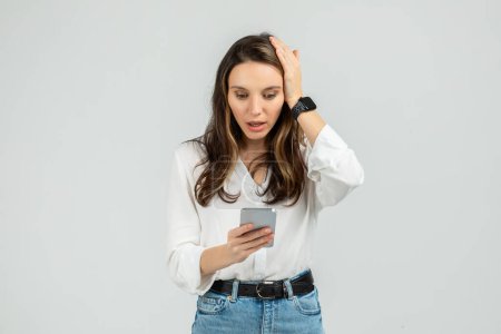 Téléchargez les photos : Surprise jeune femme en chemisier blanc et jean bleu regardant son smartphone avec une main sur la tête, montrant une réaction d'incrédulité, avec une smartwatch, sur un fond gris clair - en image libre de droit