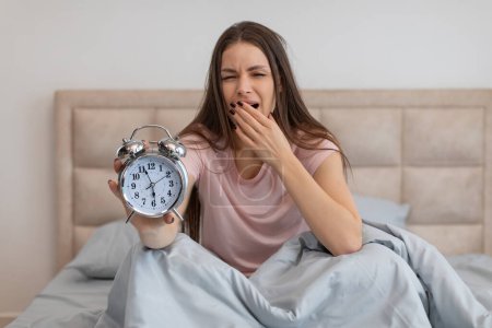 Photo for Drowsy young woman sits up in bed, stretching and yawning as she reluctantly presents an old-fashioned alarm clock, signaling the start of her day - Royalty Free Image