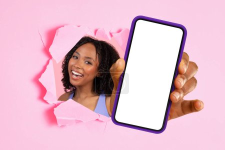 Téléchargez les photos : Femme joyeuse regardant à travers un mur en papier rose déchiré présentant un smartphone avec un écran blanc vierge, parfait pour les maquettes ou la publicité. Grande offre, annonce, site de recommandation, application - en image libre de droit