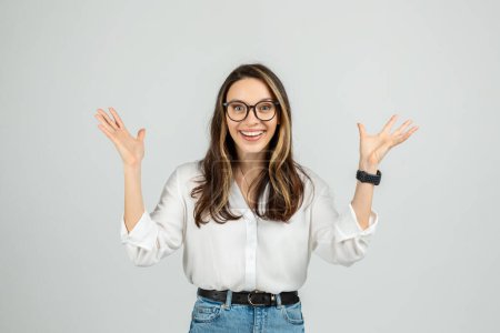 Photo for A joyful young european woman with glasses raises her hands in a shrug, wearing a white blouse and blue jeans, showing a playful expression of surprise or uncertainty, studio - Royalty Free Image