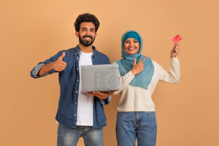 Foto de Concepto de banca online. Feliz pareja musulmana utilizando el ordenador portátil y el dedo en la tarjeta de crédito, cónyuges árabes emocionados posando sobre fondo de estudio beige, haciendo compras en línea con PC - Imagen libre de derechos