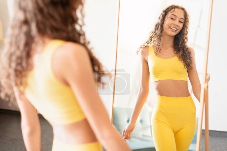 Téléchargez les photos : Adolescente positive et confiante en tenue de sport jaune sourit devant le miroir à la maison, incarnant la beauté de la jeunesse et l'engagement envers la forme physique et le bien-être. Concentration sélective - en image libre de droit