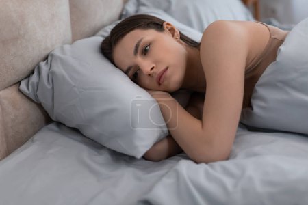Téléchargez les photos : Jeune femme couchée au lit, regardant pensivement tout en posant sa tête sur ses bras, son expression reflétant l'introspection ou le souci dans une atmosphère sereine chambre - en image libre de droit