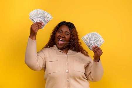 Téléchargez les photos : Lucky heureuse femme afro-américaine en surpoids avec des billets en dollars dans ses mains grimaçant sur fond de studio jaune. Joyeuse dame noire célébrant la victoire de succès à la loterie, montrant de l'argent - en image libre de droit
