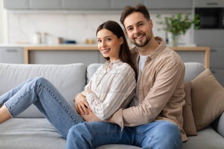 Photo for Smiling young couple sitting comfortably on gray couch, embracing and enjoying quality time together in well-lit living room with modern decor - Royalty Free Image