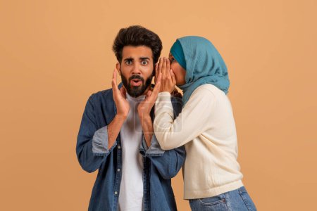 Sharing Secret. Muslimische Frau im Hijab flüstert schockierten Ehemännern ins Ohr, überraschter arabischer Mann öffnet erstaunt den Mund, während er gemeinsam vor beigem Studiohintergrund steht, Kopierraum