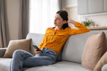 Photo for Young woman appears contemplative and concerned as she sits on grey sofa, holding her phone, looking away with head leaning on hand, in well-lit, modern living room - Royalty Free Image